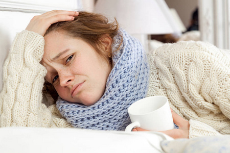 生病的妇女来杯茶。在躺在床上拿着一杯茶条蓝色围巾沮丧生病少妇的特写图像。手在头上，悲伤，看着相机