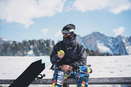 有胡子的 snowboarded 在太阳镜面具, 在滑雪胜地在山和蓝天的背景。男子休息和持有绿色苹果的手。水平模糊背景