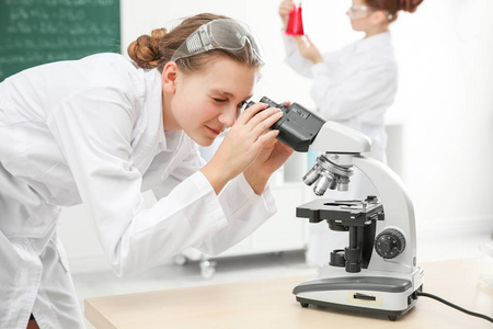 美丽的学校女孩通过显微镜在化学课