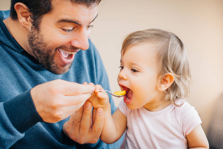 年轻快乐父亲的特写抱着勺子和喂养可爱的小女孩, 新鲜的芒果在家里