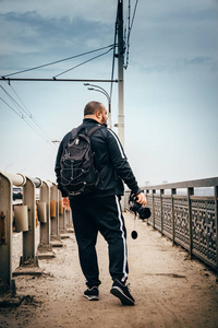 男性摄影师旅行者与背包和照相机在手步行在城市桥梁, 看法从后面, 透视, 色调