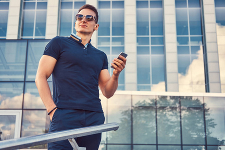 一个英俊时尚的男子与一个时髦的发型在太阳镜, 身着黑色 t恤衫和裤子, 持有智能手机, 斯坦斯在现代城市对摩天大楼