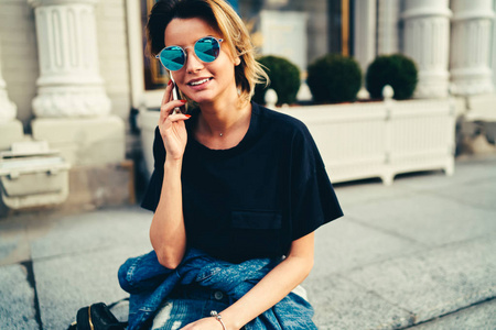 穿着黑色 t恤衫的漂亮漂亮的女士在手机上说话。时髦的蓝色太阳镜中欢快的年轻女子通过数字智能手机在城市环境中的户外交流