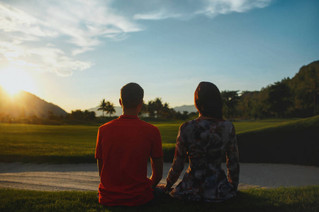 年轻夫妇坐在绿色的草地上日落。从后面查看。生活方式家庭概念