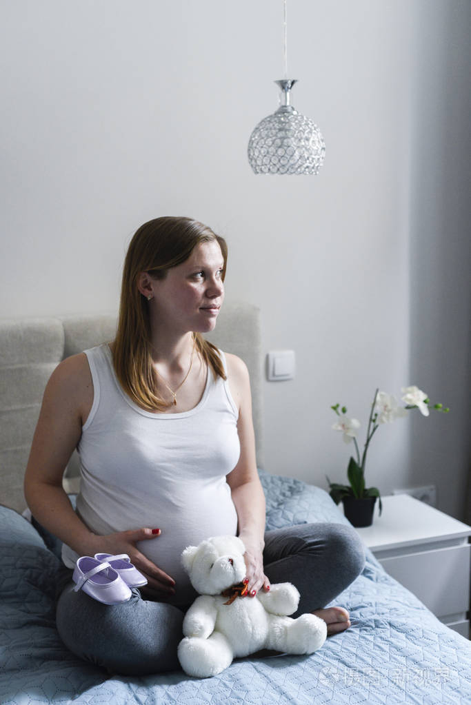 微笑的孕妇坐在床上与泰迪熊和小孩的鞋抱着她的肚子
