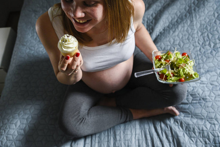 怀孕妇女一只手拿着西红柿的绿色沙拉, 吃着美味的甜蛋糕。