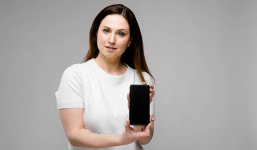 美丽的曲线黑发妇女与长的豪华头发在白色 t恤衫显示不同的表现在灰色墙上, 显示了手机的空白屏幕