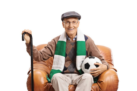 老人足球爱好者坐在真皮扶手椅上看着在白色背景下隔离的相机