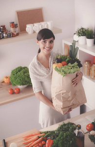 年轻女子捧着与蔬菜站在厨房里的食品杂货店购物袋