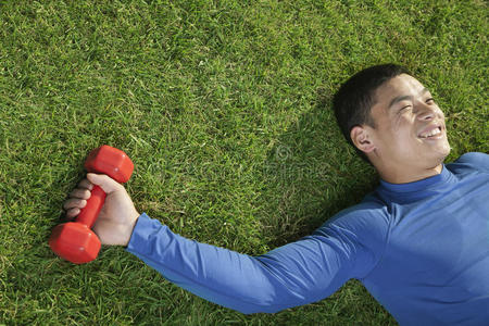 年轻快乐的运动员拿着红色的哑铃躺在草地上，从上面看
