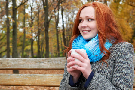 快乐的女孩杯茶坐在城市公园的长凳上的秋天
