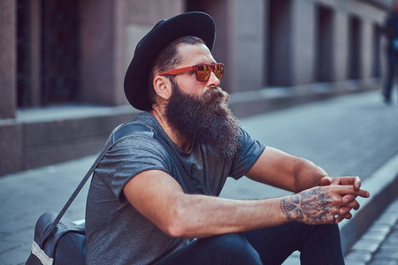 一个英俊的时髦的旅行者与一个时髦的胡子和纹身在他的胳膊穿着休闲衣服用袋子, 坐在人行道上, 休息后, 骑自行车