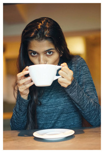 一个年轻迷人的印度亚裔妇女的肖像坐在咖啡馆的桌子上享受热咖啡。她穿着随意, 看起来舒适, 轻松和快乐