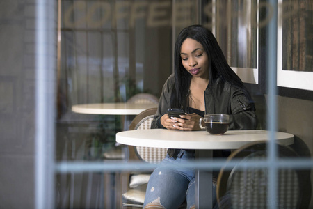 在咖啡店或人行道咖啡馆使用手机的黑人女性的窗口视图。她手持智能手机时坐着喝咖啡。