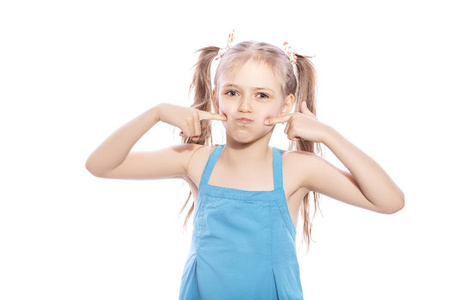 年轻的七岁的黑发女孩在蓝色礼服上的白色孤立的背景。她吹她的脸颊, 她脸上的滑稽情绪