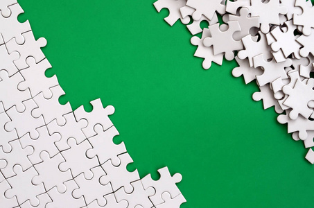 一个折叠的白色拼图拼图和一堆蓬乱拼图元素的背景下的绿色表面的碎片。带有文本空间的纹理照片