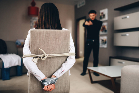 凶手在黑色的衣服与枪对他的女性受害者绑绳子和胶带的椅子上照片