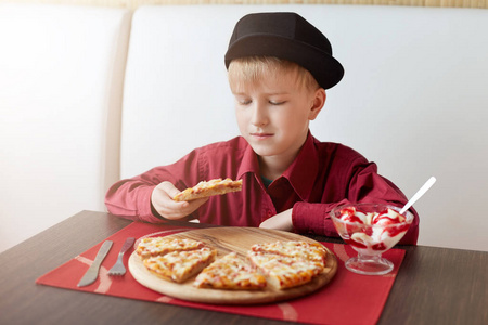 在红色的衬衫和黑色帽子坐在舒适的咖啡馆吃美味的比萨和冰激淋的时尚小孩。休息一下。休息一分钟。咖啡厅城市生活方式。休闲的儿童肖像