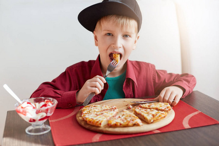 甜蜜可爱的孩子，男孩时尚帽，在一家餐馆吃比萨饼和冰激淋的红衬衫。时尚在咖啡厅吃早饭的小男孩。人和生活方式的概念