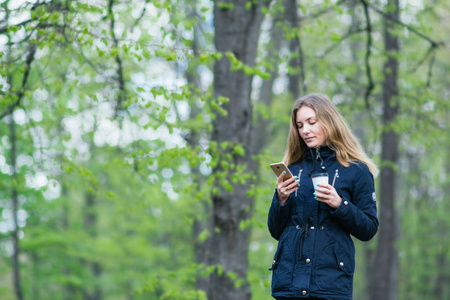 一个女孩走在早上在公园与智能手机和咖啡