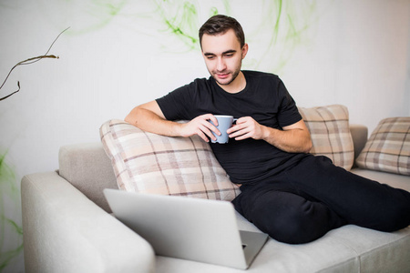 帅笑的年轻人与杯咖啡在家里使用的便携式计算机