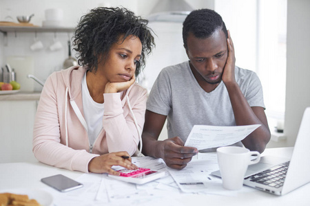 强调非裔美国人夫妇看起来沮丧 没有钱来偿还他们的债务，家庭预算管理在一起，坐在家里在厨房的桌子，与很多文件 便携式计算机和计