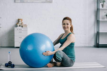 美丽的怀孕妇女坐在瑜伽垫与健身球在客厅里
