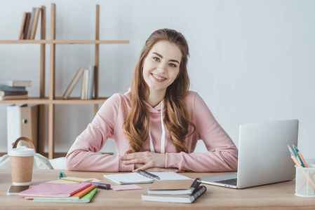 微笑的女学生坐在桌与膝上型电脑和抄写本旁