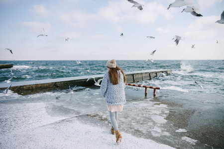 年轻女子走在冬季海滨与海鸥的背面视图