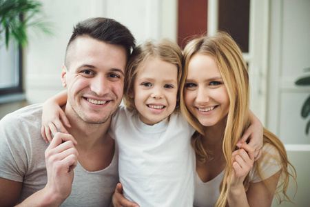 快乐的年轻漂亮的微笑的父母与一个金发碧眼的小女儿拥抱和玩耍坐在家里的沙发上。家庭概念