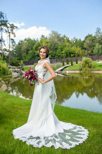 迷人的新娘在美丽的婚礼白色礼服捧着鲜花, 站在草地上, 夏日池塘边