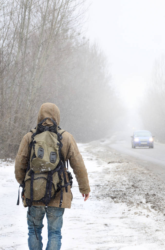 一个大背包的男人在冬天的暴风雪中沿着郊区的沥青路散步.