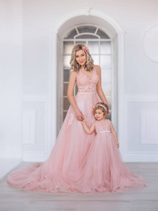 妈妈和女儿穿着豪华的粉红色连衣裙, 长着一列火车。家庭服装, 相同的礼服。背景是美丽的, 昂贵的, 白色的, 经典的内饰。艺术