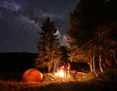 四人的朋友徒步旅行者在篝火旁温暖双手, 在夜晚异常繁星的天空和银河下, 在明亮的橙色帐篷旁休息。