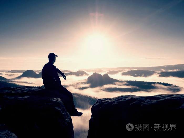 剪影的黑色和帽的独自徒步旅行者坐在悬崖上蓝色滤光