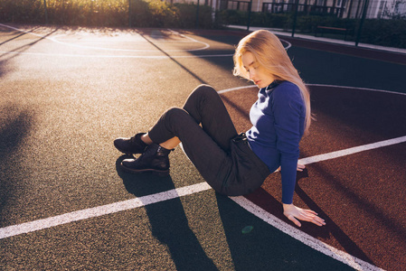 时尚的金发模特女孩穿着蓝色的毛衣坐在操场上摆在清晨的阳光照射下