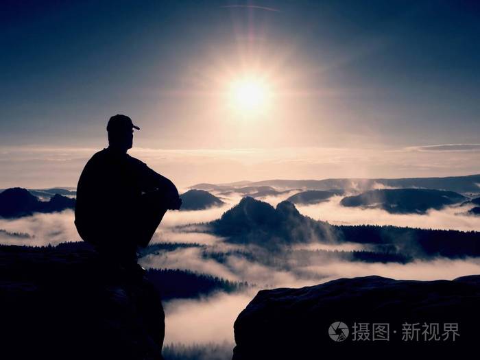 剪影的黑色和帽的独自徒步旅行者坐在悬崖上蓝色滤光