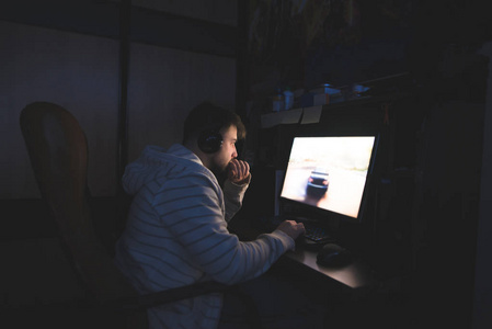 戴着耳机的那个年轻人坐在电脑边玩游戏。玩家晚上使用电脑。在电脑上赛跑。游戏玩家概念