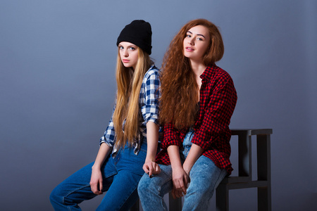 两个美丽的年轻女孩，穿着牛仔裤坐。时尚的灰色背景的女朋友画像