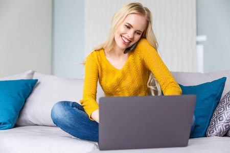 美丽的微笑年轻的金发女郎使用笔记本电脑, 并在家里的智能手机交谈