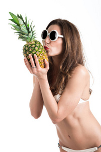 年轻健康美丽的女人白色比基尼泳装和白色太阳镜新鲜菠萝在手里的画像