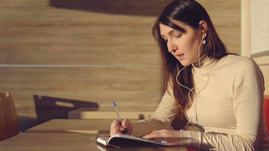 坐在咖啡馆里戴着耳机, 在笔记本上做笔记的年轻女子