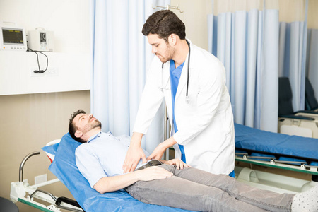 英俊的年轻医生检查在急诊室躺在床上的男性病人的腹部照片