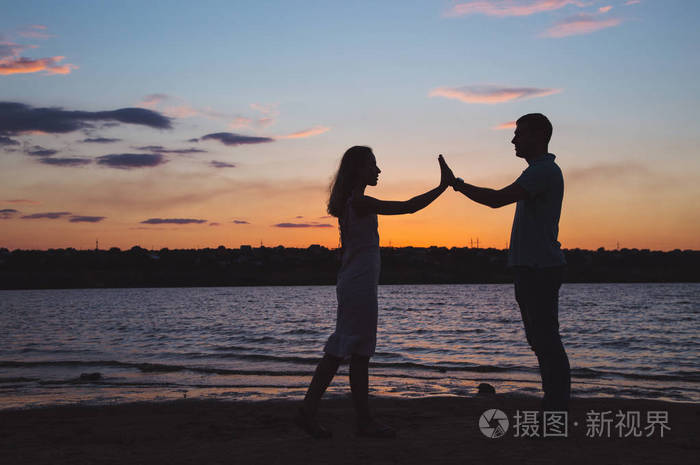年轻浪漫情侣的剪影在日落临近湖边对多云的天空牵手