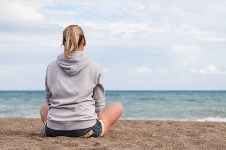 温暖晴朗的夏日。一个穿着灰色汗衫和牛仔裤短裤的女孩坐在她的背上, 在悲伤的岸边的相机, 看着大海