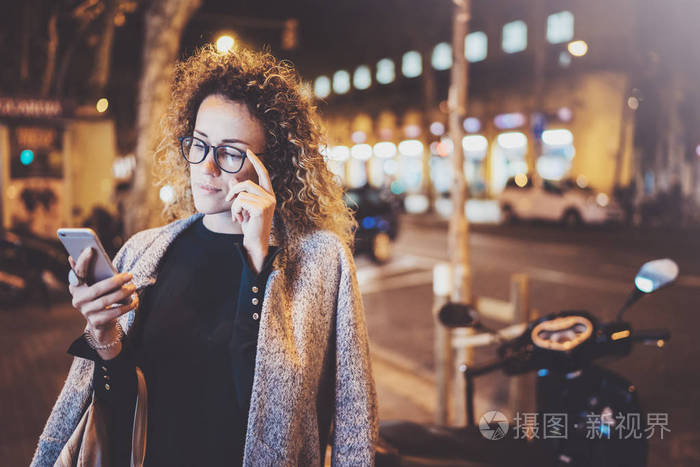 美丽的女孩时髦戴着眼镜手持智能手机手在夜间步行在欧洲的职业模糊