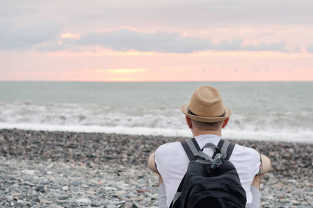 戴着帽子的年轻人带着背包坐在沙滩上。大海和日落的天空。后退视图