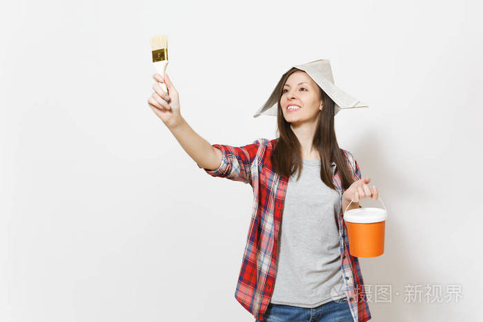 年轻的微笑美丽的女人在报纸帽子持有油漆桶和指向油漆刷在复制空间