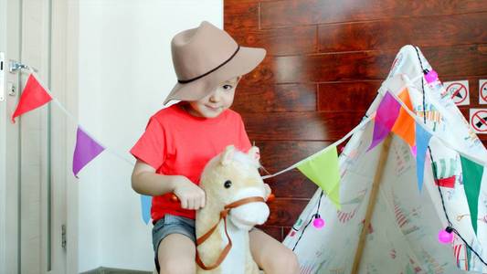小快乐男孩戴着牛仔帽骑着玩具马在游戏室里