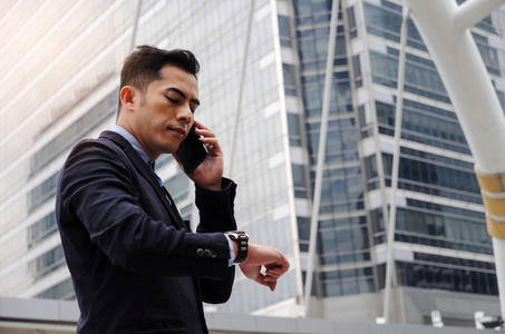 年轻精明的亚洲商人穿着现代黑色西装看着他的手表, 并拨打手机手机在城市背景, 网络连接, 技术沟通理念